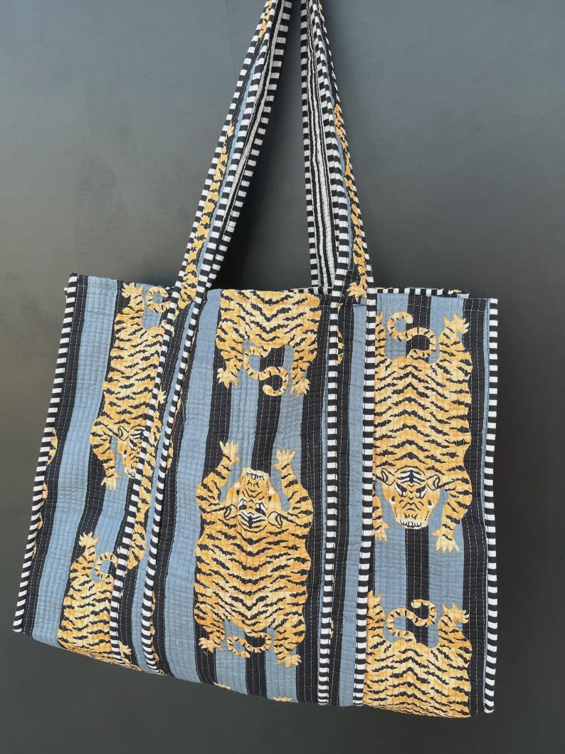 May include: A quilted tote bag with a repeating pattern of tigers in orange and black stripes against a light blue background. The bag has black and white striped handles and trim. The bag is made of fabric.