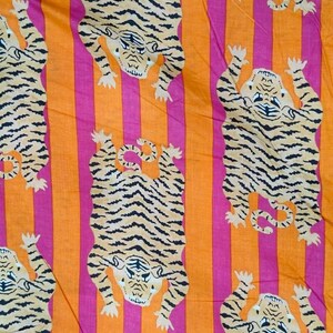 May include: Fabric with a repeating pattern of tigers in a playful pose. The tigers are tan with black stripes, set against a background of vertical stripes in orange and pink. The fabric has a bold, eye-catching design.