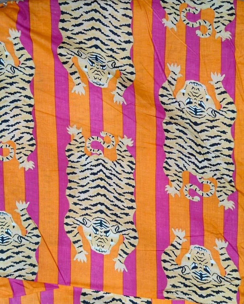 May include: Fabric with a repeating pattern of tigers in a playful pose. The tigers are tan with black stripes, set against a background of vertical stripes in orange and pink. The fabric has a bold, eye-catching design.