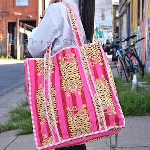 Può includere: Una grande borsa tote rosa con strisce rosse verticali e un motivo a tigre ripetuto. La borsa ha finiture e cinghie bianche e viene portata a spalla. La borsa è realizzata in materiale tessuto.