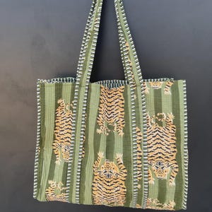 May include: Green tote bag with vertical stripes and tiger print. The bag features long handles and a rectangular shape. The tiger print is in shades of orange and black. The bag is made of a soft, quilted fabric.