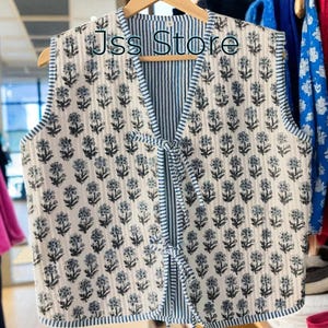 Könnte beinhalten: Eine Wendeweste mit blau-weißem Blumenmuster. Die Weste hat ein gestreiftes Innenfutter und blaue Paspeln. Sie hängt an einem Holzbügel. Der Text "Jss Store" ist auf der Weste zu sehen.