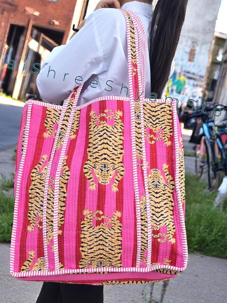May include: A pink and red striped tote bag with a repeating tiger pattern. The bag has a white trim and matching straps. The text "aishreesh yamsto" is visible on the bag.