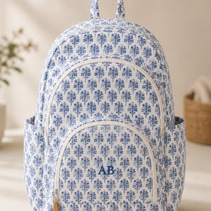 Könnte beinhalten: Ein Rucksack mit weiß-blauem Blumenmuster, ausgestattet mit einem Tragegriff, zwei Seitentaschen und einem vorderen Reißverschlussfach. Die Vordertasche ist mit den blauen Buchstaben AB bestickt und hat einen kleinen beigen Lederanhänger.