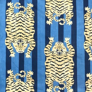 May include: Fabric with a repeating pattern of stylized tigers in cream and black against a blue and navy striped background. The tigers are depicted in a symmetrical pose, with their paws raised.