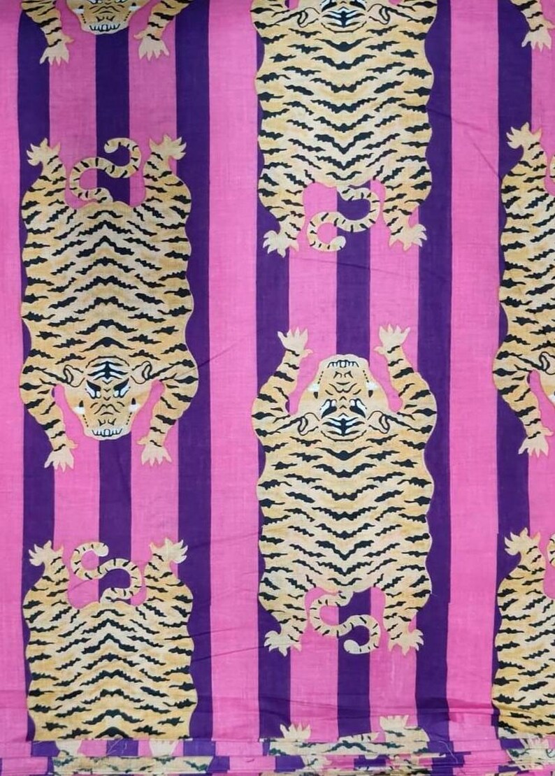 May include: Fabric with a repeating pattern of tigers in a vertical orientation. The tigers are tan with black stripes, set against a background of alternating pink and purple vertical stripes. The tigers are depicted with their paws up.