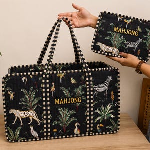 Op de afbeelding: Zwarte tote bag en bijpassende etui met een tropische print met zebra's, luipaarden, vogels en bladeren. Het woord "MAHJONG" is op beide items geborduurd. De tote bag heeft zwart-wit geblokte handvatten en afwerking.