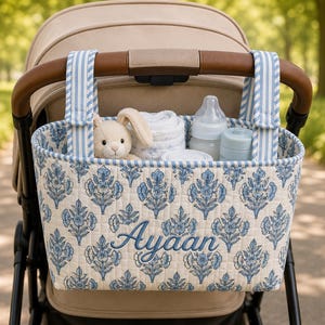 May include: A quilted fabric stroller organizer featuring a blue and white floral pattern with the name Ayaan embroidered in blue script. It has two blue and white striped hanging straps and contains a plush rabbit, a baby bottle, and cloth.