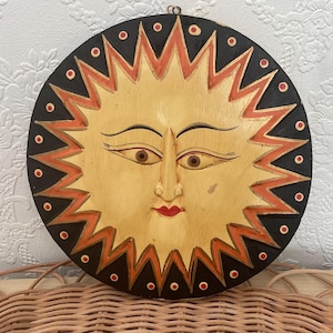 Peut inclure: Décoration murale en bois en forme de soleil. Le soleil a un visage jaune clair, des yeux bruns et une bouche rouge. Les rayons du soleil sont orange et noirs, avec une bordure noire. Une petite boucle métallique est en haut pour accrocher.