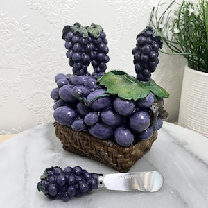 Peut inclure: Un panier tressé marron rempli de raisins violets et de feuilles vertes, accompagné d'un couteau à fromage assorti avec un manche en forme de grappe. La lame métallique du couteau porte l'inscription STAINLESS, indiquant sa composition.