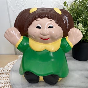 May include: A vintage ceramic doll figurine with brown hair, yellow accents, and a green dress. The doll has a smiling face with red lips and is posed with arms outstretched. The figurine is on a marble surface.