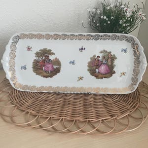 Peut inclure: Un plateau de service rectangulaire en porcelaine blanche avec des bordures et des poignées dorées. Le plateau présente deux scènes ovales de couples en costumes d'époque, entourées d'accents floraux. Le plateau est posé sur un dessous de plat rond et tissé.