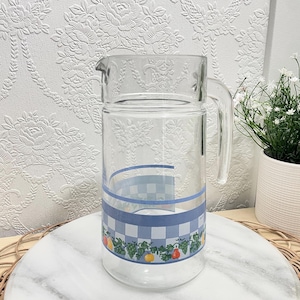 Peut inclure: Une carafe en verre transparent ornée d'une bande à carreaux bleus et blancs au centre, agrémentée d'une bordure décorative de feuilles vertes et de divers fruits colorés, dont des formes rouges, jaunes et orange près de la base.