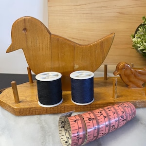 Peut inclure: Porte-fil en bois en forme d'oiseau avec deux bobines de fil noir et une petite figurine d'oiseau en bois. Un mètre ruban rose est enroulé devant le porte-fil. Le support est sur une surface en marbre blanc.