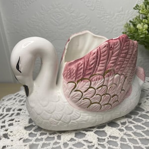 Peut inclure: Un pot de fleurs en forme de cygne en céramique, principalement blanc avec des accents roses et dorés. Les ailes du cygne sont rose clair avec des détails dorés, et le bec et les yeux sont noirs. Le pot est conçu pour contenir de petites plantes ou fleurs.