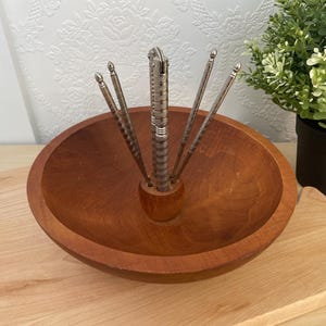 May include: A wooden nut bowl with a built-in nutcracker and four nut picks. The bowl is round and made of a warm-toned wood. The nutcracker and picks are silver-coloured metal. A small potted plant is in the background.