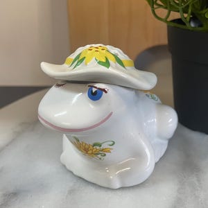 May include: A white ceramic frog ornament wearing a hat decorated with yellow sunflowers. The frog has blue eyes and a pink smile. The ornament is on a marble surface, with a black potted plant in the background.