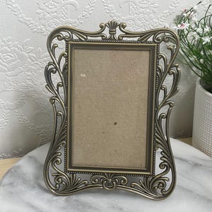 Peut inclure: Cadre photo orné de couleur bronze avec une ouverture rectangulaire. Le cadre présente des volutes complexes et un dessus décoratif. Le cadre est présenté sur une surface semblable à du marbre.