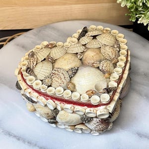 Peut inclure: Une boîte en forme de cœur décorée d'un assortiment de coquillages naturels dans des tons beige, crème et marron. Le couvercle et les côtés sont recouverts de divers types de coquillages, dont de petits coquillages circulaires en bordure.