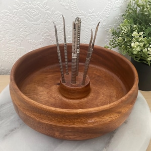 Peut inclure: Un bol à noix en bois avec un support central contenant plusieurs cure-noix argentés. Le bol est rond et en bois aux tons chauds. Le bol repose sur une surface semblable à du marbre blanc et gris.
