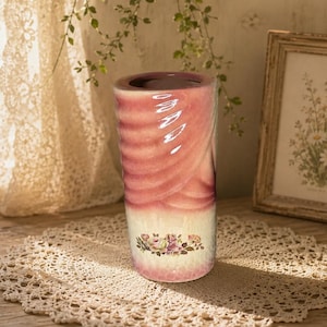 Peut inclure: Un vase cylindrique avec un motif tourbillonnant rose et blanc. Le bas du vase présente un motif floral. Il est posé sur un napperon en dentelle, avec un cadre photo et un petit vase de fleurs en arrière-plan. Le vase mesure environ 20 cm de haut.