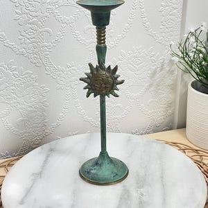 May include: A tall, slender candlestick holder with a weathered, verdigris-green patina finish. The stem features a central, circular sun face emblem in a bronze tone, positioned below a ribbed neck and a wide, circular top candle cup.