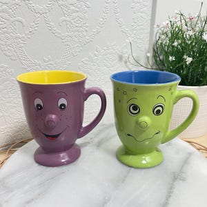 Peut inclure: Deux tasses en céramique avec des traits de visage en relief. La tasse de gauche est violette avec un intérieur jaune et un visage souriant. La tasse de droite est vert citron avec un intérieur bleu, une bouche ondulée et des étoiles.