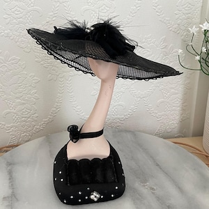 May include: A black hat display stand featuring a black hat with a mesh brim and feather accents. The stand has a black base with a decorative bow and rhinestone details. The hat is displayed on a slender, pale-toned neck.