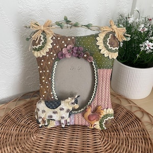 May include: A decorative photo frame with a farm theme. The frame features a cow and a rooster, with sunflower and floral accents. The frame is made of various materials and has an oval opening for a photo. The frame is approximately 25 cm tall.