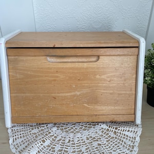 May include: A wooden bread bin with a light brown finish and a white frame. The bin has a handle on the front and a hinged lid. The bread bin is on a white lace doily.