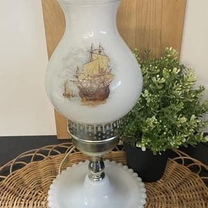 May include: A vintage white glass lamp with a ruffled top and base. The lamp features a detailed illustration of a sailing ship on the glass shade. The base is silver-toned, and the lamp is on a woven surface.