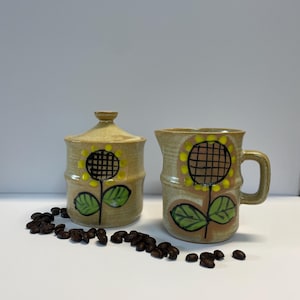May include: A ceramic sugar bowl with a lid and a creamer, both decorated with a sunflower design. The pieces are beige with yellow, green, and brown accents. Coffee beans are scattered around the base of the items.