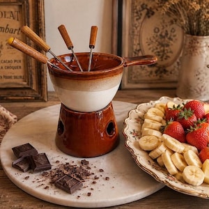 Peut inclure: Un caquelon à fondue en céramique avec un vernis bicolore marron et crème, doté d'un socle assorti avec des ouvertures, d'une poignée latérale et de quatre fourchettes en métal à manche en bois, accompagné de carrés de chocolat et de fruits.