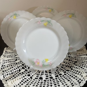 Peut inclure: Quatre assiettes blanches à bord festonné avec un motif floral. Chaque assiette présente un arrangement délicat de fleurs roses et jaunes avec des tiges vertes. Les assiettes sont présentées sur une nappe en crochet crème.