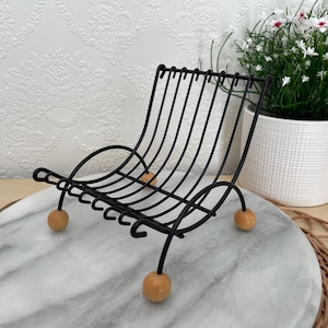 May include: A black metal wire chair-shaped rack with wooden ball feet. The rack has a curved back and seat, and is designed to hold items. The rack is sitting on a marble surface.