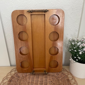 Peut inclure: Plateau de service en bois avec huit découpes circulaires pour verres ou bouteilles. Le plateau a une forme rectangulaire avec une section centrale et une poignée en haut. Le bois a une teinte naturelle et chaude, avec des ferrures couleur laiton.