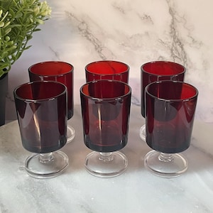 Peut inclure: Six gobelets vintage en verre teinté rouge avec des tiges transparentes. Les verres ont une forme cylindrique avec un bord évasé. Les gobelets sont disposés sur une surface blanche, avec une plante dans un pot noir en arrière-plan.