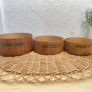 Peut inclure: Trois bols ronds en bois avec une finition en grain de bois brun clair. Les bols portent des inscriptions en noir indiquant POTATO CHIPS, PRETZELS et PEANUTS. Les bols sont disposés en rangée, diminuant de taille de gauche à droite.