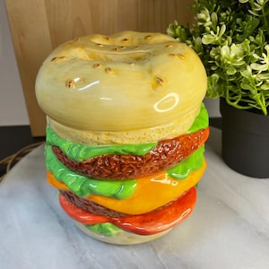May include: A ceramic burger-shaped container with a yellow bun, green lettuce, a brown patty, orange cheese, and red tomato. The container is designed to resemble a classic hamburger. The container is a decorative kitchen item.