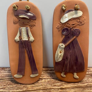 Peut inclure: Deux plaques murales de couleur terre cuite représentant des enfants en relief. L'une représente un garçon en casquette et salopette, l'autre une fille en robe et chapeau, tenant un sac. Les deux ont des boucles de suspension en cuir.