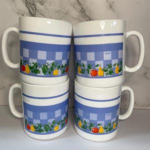 Peut inclure: Quatre tasses en céramique blanche empilées. Chaque tasse présente une bande à carreaux bleus et une bordure décorative avec des illustrations de fruits et de vignes. Les tasses ont de grandes anses incurvées.