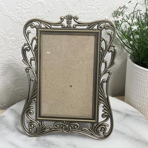 May include: A rectangular picture frame with an ornate, antique bronze-coloured metal border featuring intricate scrollwork and filigree detailing. The frame has a central rectangular opening with a plain, light brown backing insert.