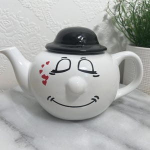 May include: A white ceramic teapot with a cartoon face design. The teapot has a black hat, closed eyes, a nose, and a smiling mouth. Red heart accents are on the side. It has a handle and a spout.