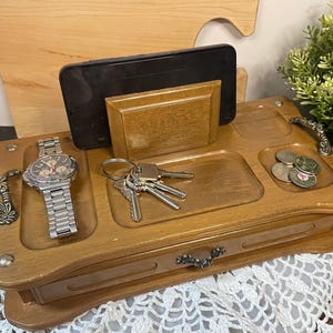 May include: Wooden organiser with compartments for a watch, keys, coins, and a smartphone. The organiser has a drawer and decorative handles. A silver watch with a metal band is in the left compartment. The organiser is made of light brown wood.