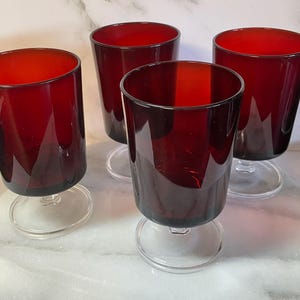 Peut inclure: Quatre verres à vin rouge rubis avec des pieds et des bases transparents. Les verres ont une forme cylindrique et sont disposés sur une surface en marbre blanc. Les verres sont vides.