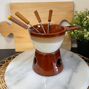 Peut inclure: Un service à fondue vintage avec un pot en céramique marron et blanc, une base assortie et plusieurs fourchettes à fondue. Le pot a une poignée et est rempli de fourchettes. L'ensemble est sur une surface en marbre.