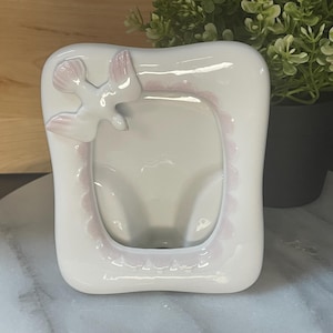 May include: A white ceramic photo frame featuring a pink dove and scalloped edging. The frame has a glossy finish and a rectangular shape with rounded edges. A small potted plant is in the background.