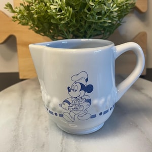 Peut inclure: Crémier en céramique blanche avec intérieur et anse bleus. Le crémier présente une illustration bleue de Mickey Mouse avec un chapeau et un tablier de chef. Un motif pointillé bleu encercle la base du crémier.