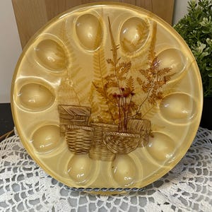 Peut inclure: Un plat de service vintage, rond, de couleur jaune, avec 12 empreintes en forme d'œuf. Le centre présente un motif botanique avec des fougères et des illustrations de paniers. Le plat est soutenu par un support en fil métallique.