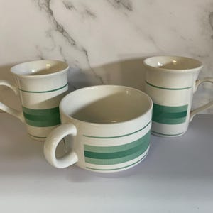 Peut inclure: Trois tasses en céramique blanche et un bol avec des rayures horizontales vertes. Les tasses ont des anses et une forme cylindrique. Le bol est rond avec une anse. Les rayures varient en nuances de vert.